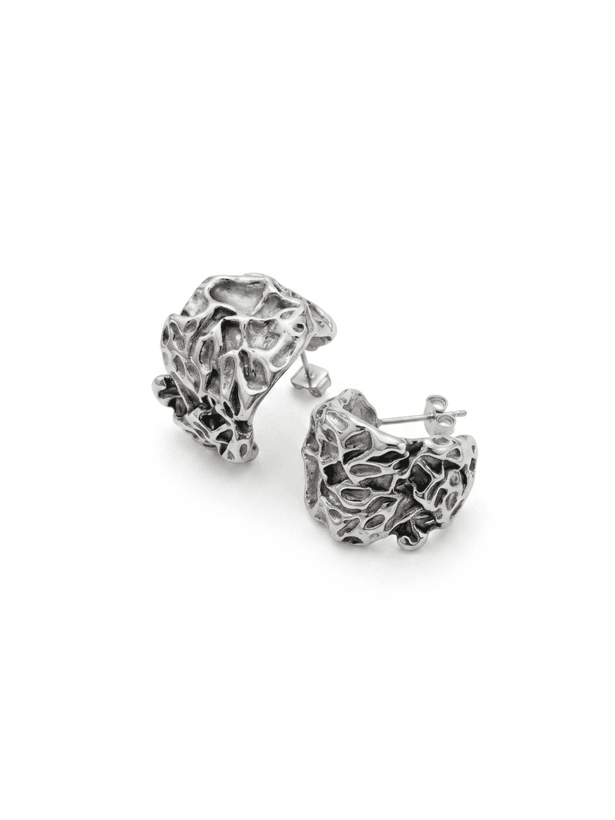 Pair of wide silver half-hoop earrings with a pierced, eroded lattice surface and oxidised recesses, on a pale grey backdrop.