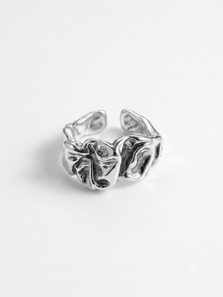 Open silver ring of crushed, folded metal with darkened recesses and polished raised edges, on a pale grey backdrop.